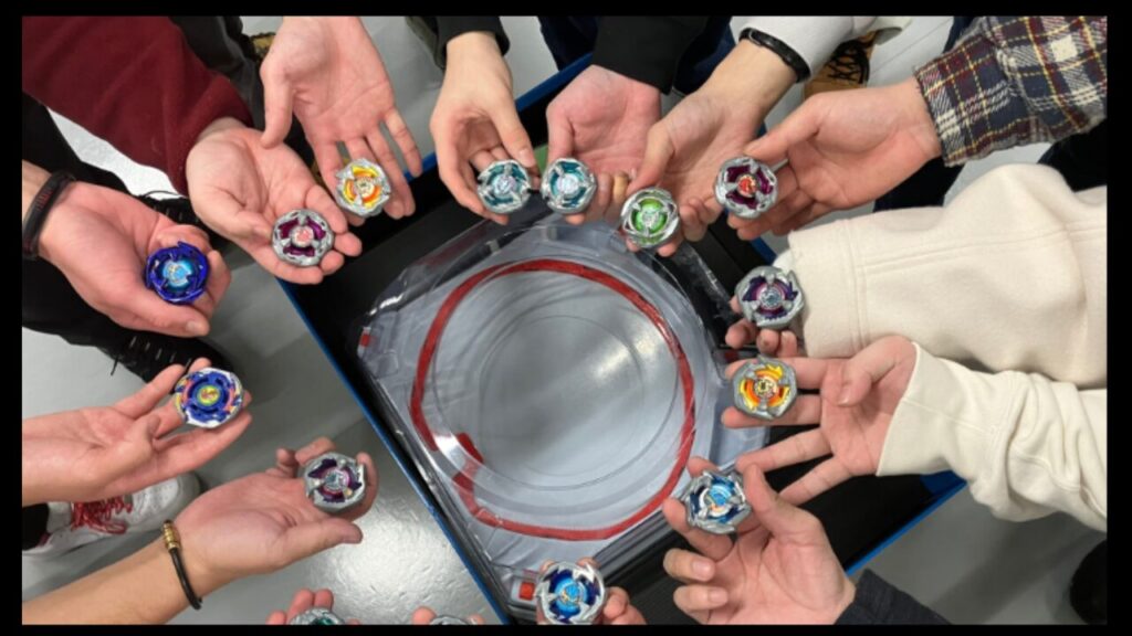 ベイブレードXのスタジアムを囲み、参加者がそれぞれのベイを手に集まる様子